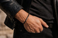 May include: A beaded bracelet featuring small, round, matte black beads and a single, textured, cylindrical metallic accent piece in a dark gunmetal finish, worn on a wrist with a black leather jacket and a black shirt.