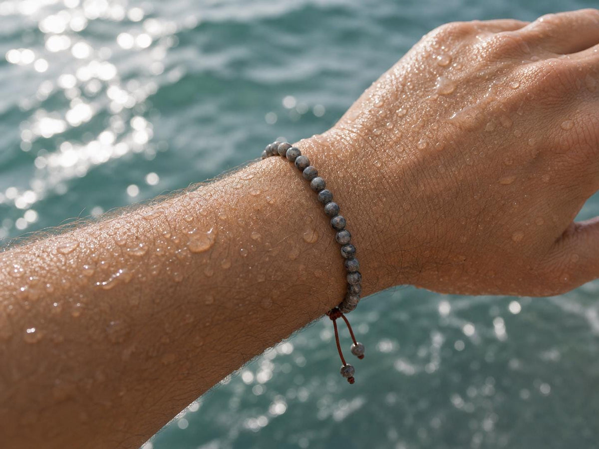 May include: A beaded bracelet featuring small, round, dark gray textured beads arranged in a single strand. The bracelet includes a dark brown cord closure with two dangling ends, each finished with a single matching gray bead at the tip.