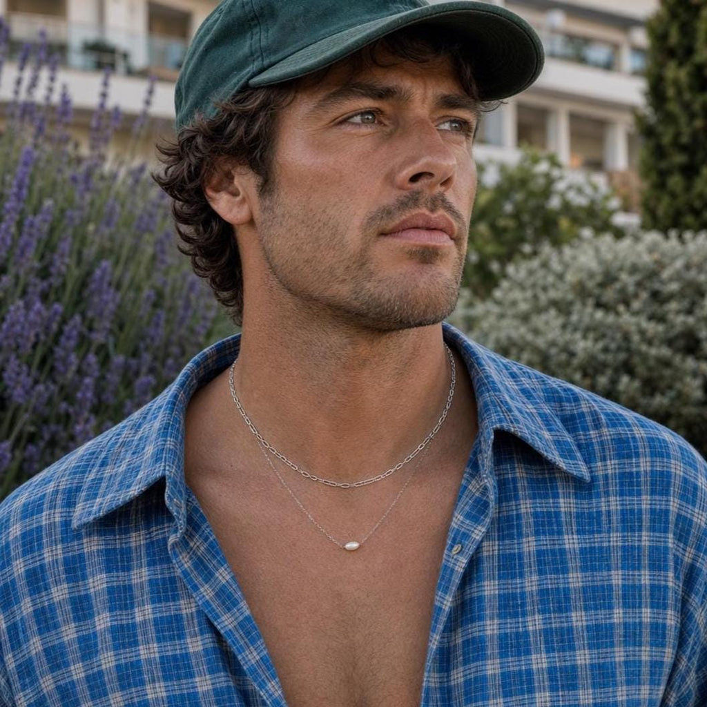 May include: A layered silver necklace featuring a delicate chain with a small, oval-shaped white pearl pendant and a second, slightly thicker silver chain with elongated oval links, worn around the neck with a blue and white plaid button-down shirt.