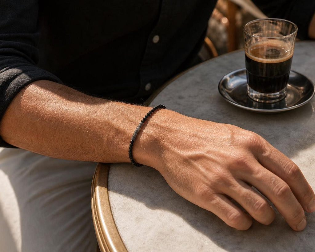 May include: A beaded bracelet featuring small, round, matte black beads and a single, cylindrical, textured dark gray metal accent bead. The bracelet is worn on the wrist, showcasing a simple, uniform design with a consistent bead size throughout.